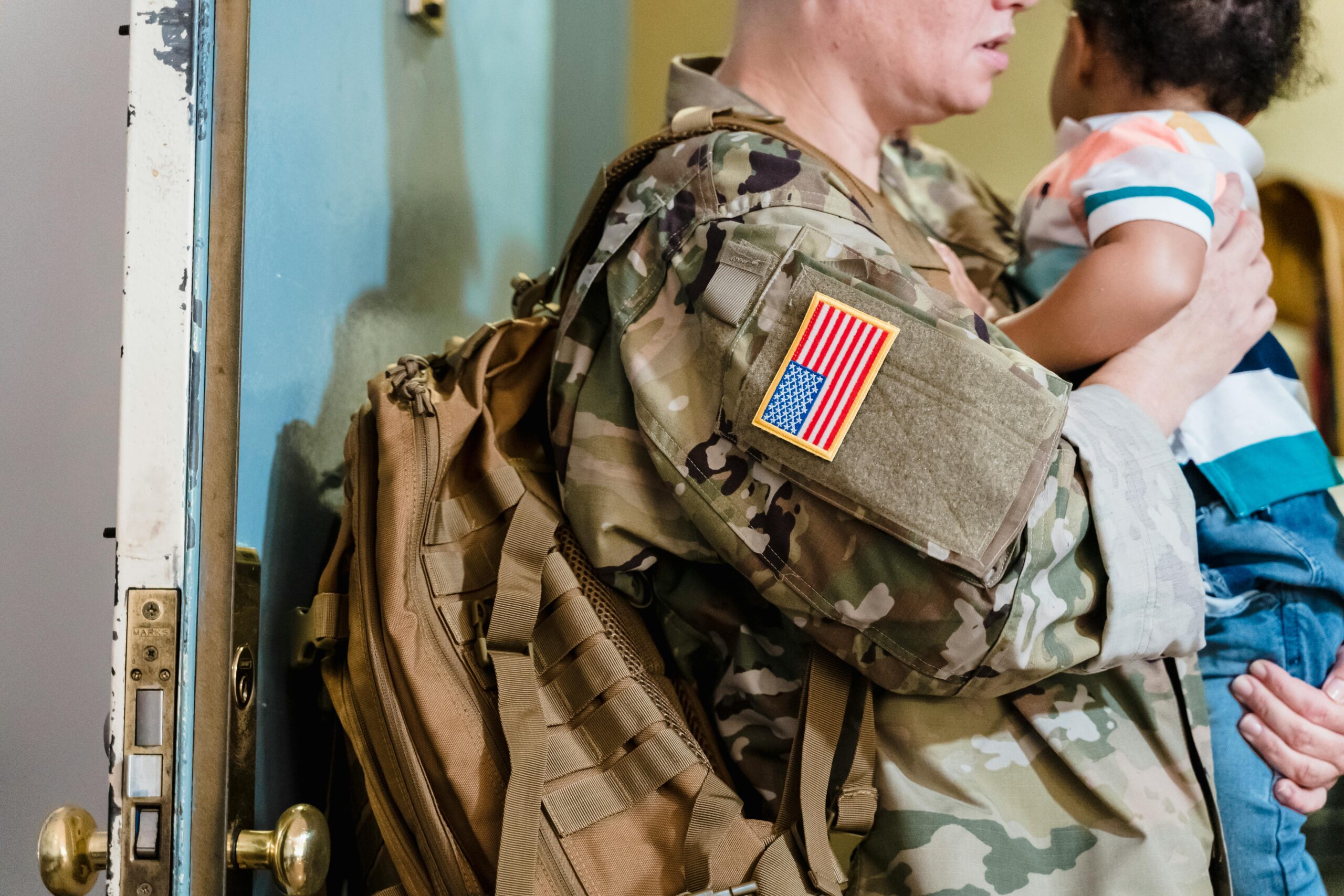 A soldier in camouflage uniform embraces a young child indoors, showing family warmth.